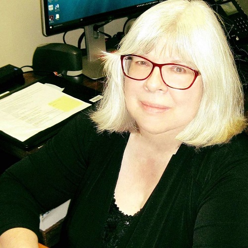 image of Mickie Hayes, platinum hair, red glasses, black jacket, sitting at a desk, author of this website, Learning Disability Specialist who supports all students with disabilities